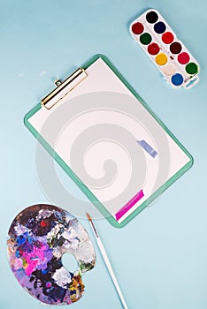 Clipboard with blank paper sheets,multicolored watercolor paints in box, paint brushes and palette on blue table. Top