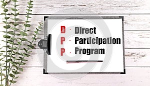 Clipboard with text direct participation program and pen on white wooden background with green plant