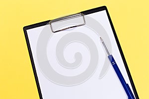 Clipboard with white sheet and pen on a yellow background. View from above. space for text