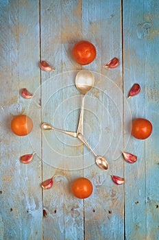 Clock arranged from tomatoes, garlic and spoons. Textured abstract clock face showing 5. Blue background