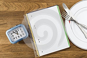 Clock, notebook and dish with cutlery on a wooden background