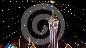 Clock tower decorated with Christmas lights, bulb garlands on city square New Year market. Camera flies around