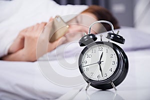 Clock with woman using her smartphone on bed