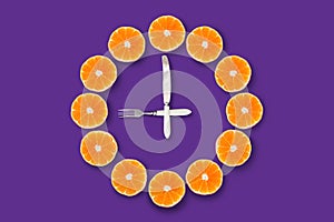 Clocks made of half cut tangerines with arrows made of fork and knife that shows 9 o`clock on a purple background.