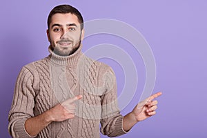 Cloe up portrait of handsome man dresses beige sweater looks curiously directly at camera, pointing with index fingers aside,