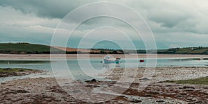 Clonakilty Bay at low tide on a cloudy day. A small blue fishing boat is anchored. Open seabed, silt and algae. Picturesque
