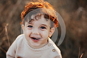 Close portrait of a sweet smiling toddler baby boy in autumn park on sunset. Curly hair boy