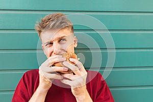 Close portrait of a young man who eats a burger and looking up against the backdrop of a turquoise wall