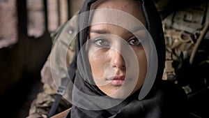Close portrait of young muslim woman in hijab looking at camera, armed soldier with gun standing behind woman, military