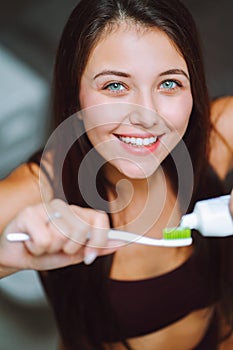 Close is portrait of young woman holding toothpaste and a toothbrush. Feemale look into camera and smile with a snow