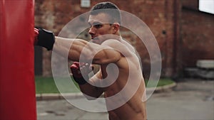 Close side view of muscular male boxer practicing punches and kicks with a punching bag being outdoors