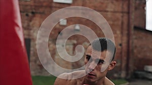 Close side view of muscular male boxer practicing punches and kicks with a punching bag being outdoors