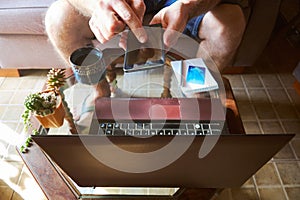 Close up from above of young man in casual clothes using mobile phone while sitting on the couch at home with laptop and credit