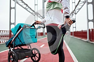 Close up of active mother in sportswear warming up her legs, standing on the bridge with a stroller on a cloudy day in