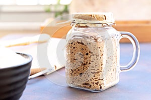 Close-up of active sourdough starter in glass jar. Rye leaven for bread and kitchen utensil on rustic background. Selective focus