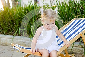 Close up Adorable blondy baby girl in dress or toddler child sitting and relaxing on a sunbed or a deckchair in the city park recr