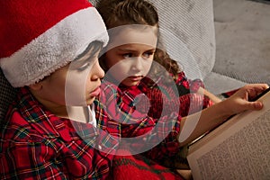 Close-up of adorable Caucasian kids, brother and sister, boy in Santa hat and beautiful girl sitting on the couch and reading