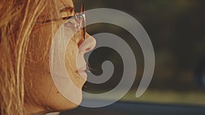 Close-up of adorable young blonde face wearing stylish sunglasses driving her car at sunset. Pretty girl listening to