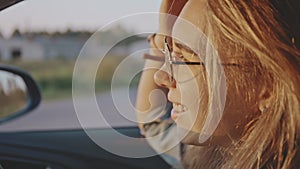 Close-up of adorable young blonde portrait wearing stylish sunglasses driving her car at sunset. Pretty girl listening