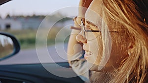 Close-up of adorable young blonde portrait wearing stylish sunglasses driving her car at sunset. Pretty girl listening