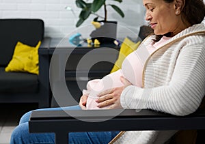 Close-up of an adult pregnant woman smiling, touching her belly, feeling baby movements, sitting relaxed on sofa indoors