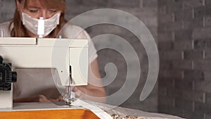 Close up of adult woman stitching curtain, using sewing machine. Young seamstress in medical mask working on sewing