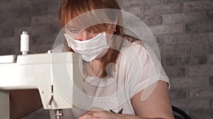 Close up of adult woman stitching curtain, using sewing machine. Young seamstress in medical mask working on sewing