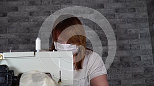 Close up of adult woman stitching curtain, using sewing machine. Young seamstress in medical mask working on sewing