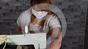 Close up of adult woman stitching curtain, using sewing machine. Young seamstress in medical mask working on sewing