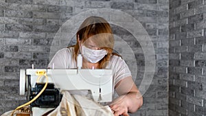 Close up of adult woman stitching curtain, using sewing machine. Young seamstress in medical mask working on sewing