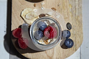Close-up with aerial view of a glass jar with plain yogurt, blueberries, muesli and raspberries on a wooden board