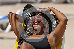 Close-up of an African-American woman on the beach lying on a deck chair wearing a black hat and sunglasses holdin her hat with