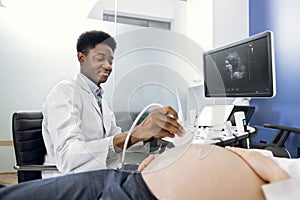 Close-up of African man doctor sonographer scanning young pregnant woman& x27;s belly with ultrasonic transducer doing