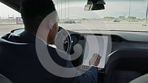 Close up African man in suit sitting on driver's seat of electric car and touching dashboard with finger. Young man