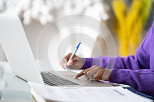 Close up of Afro-American woman employee or student using laptop, touches the touchpad with your finger, makes notes with pen