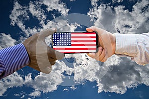 Close up of afroamerican and caucasian white hands holding the phone with US flag in front of clouds. America above all