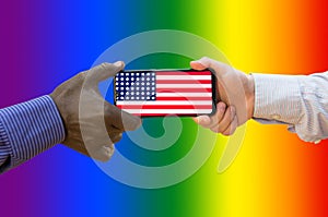 Close up of afroamerican and caucasian white hands holding the phone with US flag in front of a rainbow. LGBT community