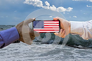 Close up of afroamerican and caucasian white hands holding the phone with US flag in front of the sea. America above all