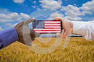 Close up of afroamerican and caucasian white hands holding the phone with US flag in front of a wheat field. America above all