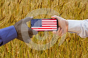 Close up of afroamerican and caucasian white hands holding the phone with US flag in front of a wheat field. America above all