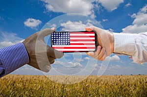 Close up of afroamerican and caucasian white hands holding the phone with US flag in front of a wheat field. America above all