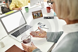 Close up of aged woman, senior intern holding, taking pills while using laptop, sitting at desk, working in modern