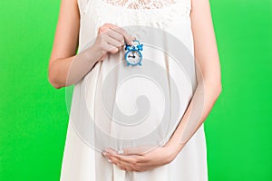 Close up of alarm clock against pregnant woman`s belly in white dress at green background. Copy space. Prenatal concept