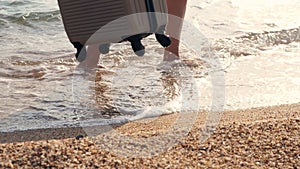 Close-up, along sandy beach, in foam of surf, female legs are walking. woman carries a travel suitcase. travel concept