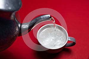 Close-up of aluminum teapot pouring water in ceramic mug on background of red color.