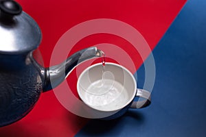 Close-up of aluminum teapot pouring water in ceramic mug, on two backgrounds of red and blue colors. Top view.