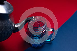 Close-up of aluminum teapot pouring water in glass mug for juice, on two backgrounds of red and blue colors. Top view.