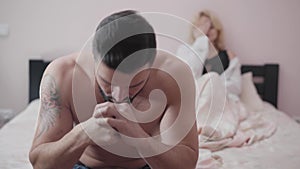 Close-up of angry Caucasian man sitting on bed. Blurred blond woman lying at the background. Wife cheating her husband