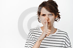 Close up of angry girl hushing, frowning and say shh, press finger to lips, taboo gesture, standing against white