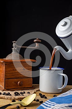 Close-up of antique coffee maker serving coffee in white cup with old coffee grinder, coffee beans and spoon on wooden table and b